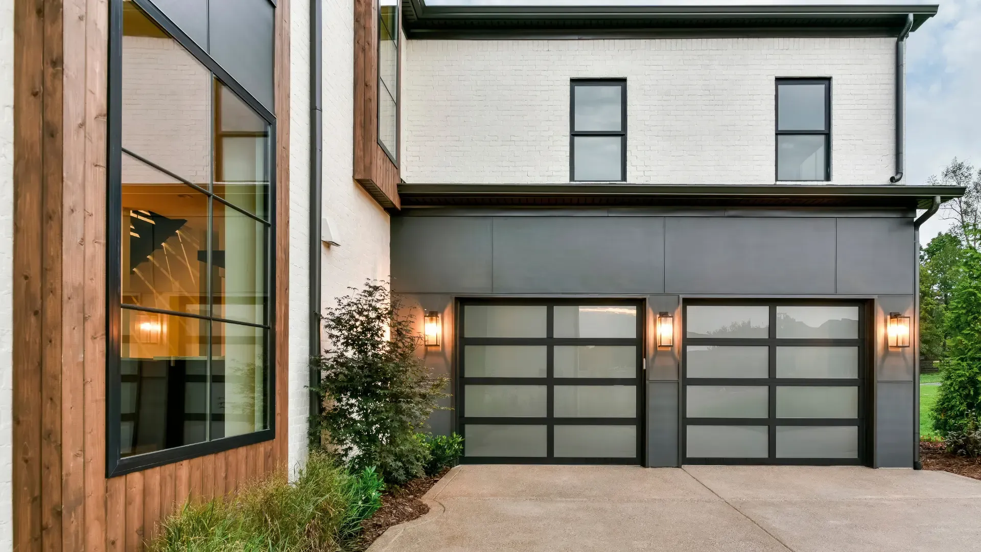 A Clopay Avante Collection full view sectional garage door with a black aluminum frame and illuminated glass panels, installed on a modern two-story home.